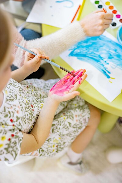 A child is painting their hand with pink paint using a brush, while another person paints a blue design on paper nearby, surrounded by watercolor paints on a table.