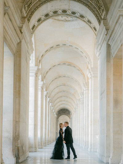 a couple in black outfits walking away from the camera and leaning towards each other for a kiss with beautiful arches at the louvre