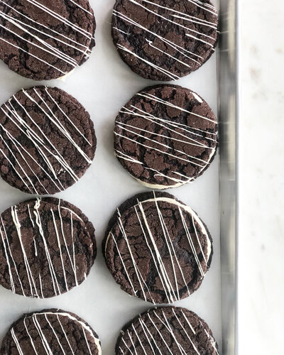 Black cocoa cookie sandwiches drizzled in white chocolate on a tray.