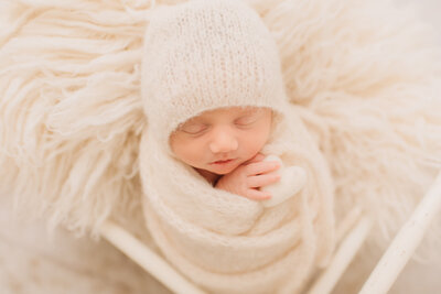 A close-up of a baby girl, wrapped in white, holding a white heart prop at her newborn session