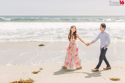 Engaged couple walk along Santa Monica State Beach while holding hands