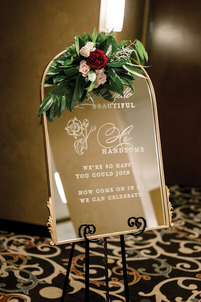 Gold-framed mirror Welcome sign, adorned with burgundy and blush floral accents, for a 50th wedding anniversary celebration.