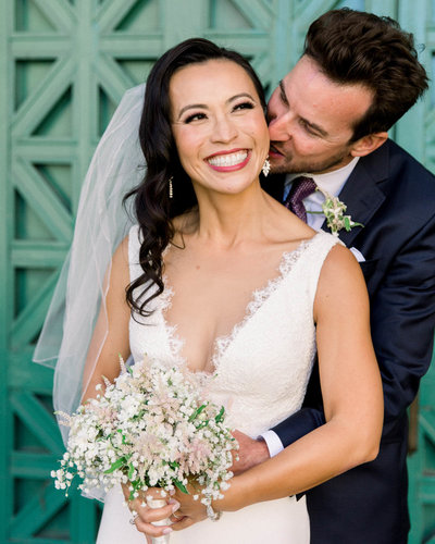 Groom stands behind Bride kissing her left cheek as she smiles holding bouquet