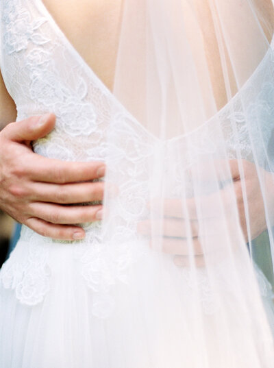 Groom hands wrap around brides waist, wearing wedding gown and veil