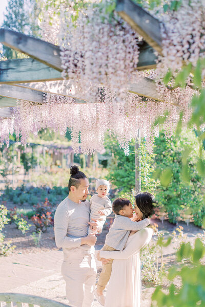 A mother and father hold their two sons while standing under purple wisteria at Brookside Gardens photographed by Family Photographer Northern Virginia Marie Elizabeth Photography