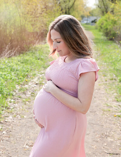 A glowing mom-to-be gently holding her belly during an outdoor photography session in Rochester, NY, surrounded by lush greenery and soft sunlight, capturing the beauty of motherhood.