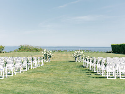 Outdoor empty wedding ceremony space in the grass on the water with white chairs and floral arrangements down the aisle and on the altar