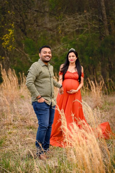 expectant couple in a field with dad as primary focus wearing a green shirt and jeans and mom holding his hand behind him in a rust colored dress and crown