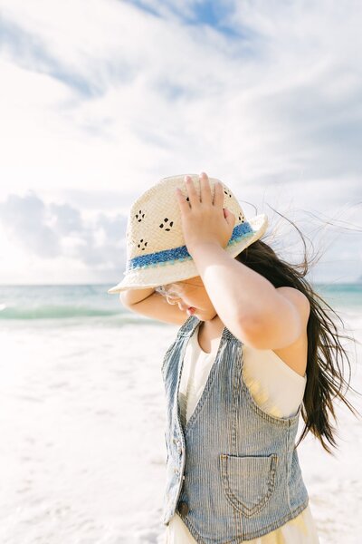 A girl holds her hat on her head as the wind blows through her hair on a beach in Hawaii.