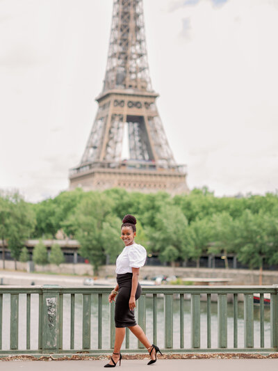 a black woman in a white top and black skirt walking along the Bir Hakeim bridge in Paris with the eiffel tower behind her