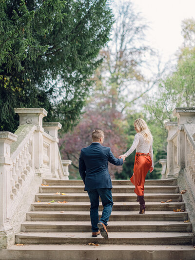 a blonde woman in an orange skirt and white top walking up some steps and holding hands with her husband who is in a blue suit and jeans