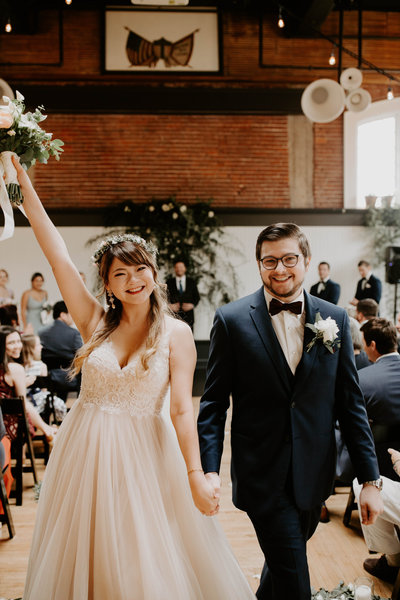 Bride holds her bouquet up and smiles while walking out of her reception hand in hand with her groom at The Evergreen in Portland, Oregon
