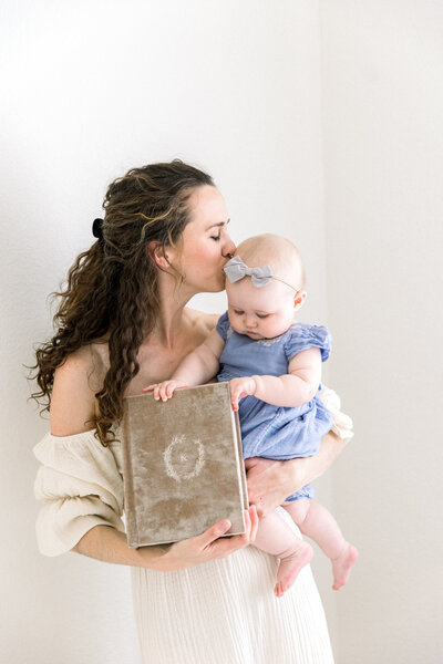 Image of Newborn Photographer Sacramento Kelsey Krall holding heirloom album and her baby girl and kissing her head