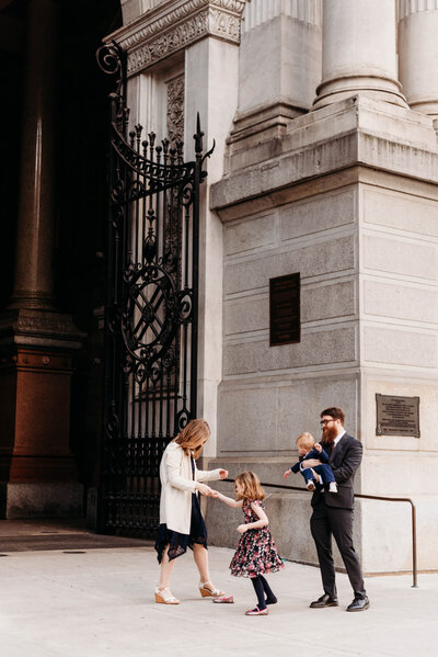 Mom dancing with her daughter, while dad holds young son and looks on.  Family stands in front of the arch at City Hall during their Philadelphia Family Photography session