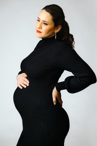 Maternity portrait of a woman in a black dress and denim jacket, leaning against a tree outside her Chestnut Hill home with her eyes closed.