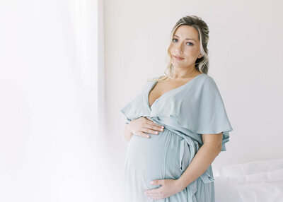 Soon to be mother in a flowy sky blue maternity dress cradling her baby bump.