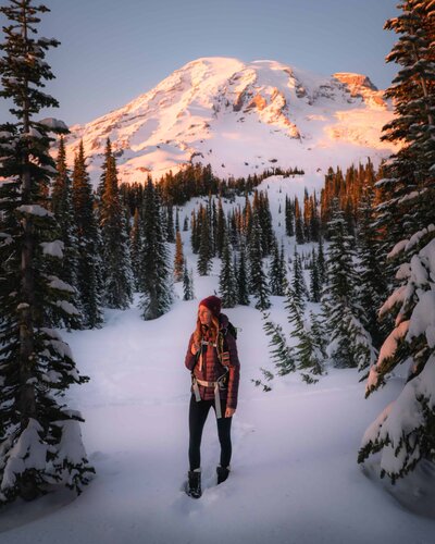 Woman walking in snow surrounded by pine trees with snowy mountain behind