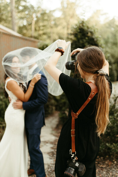 Wedding photographer taking pictures of a bride and groom while holding the bride's veil in front of camera
