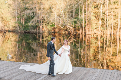 A smiling bride holding the hand of her new husband on the Neuse River during the fall by JoLynn Photography