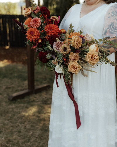 Bridal bouquet in fall tones with roses, zinnias and more, held by woman in white dress.