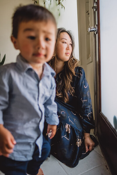 Asian mother gazes out of window while her toddler approaches camera