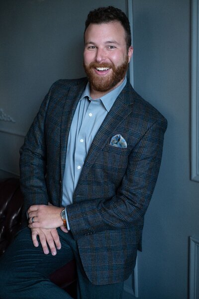 A headshot of Greenville SC wedding photographer, Zack Bradley wearing a brown tweed suit with blue and green accents including a pocket square, a brown skagen watch, and a gray shirt.