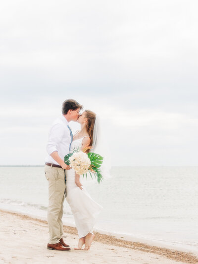 A bride and groom kiss on a sandy beach as the ocean shimmers behind them. The bride holds a bouquet of white flowers and greenery while the groom wears a white shirt and khaki pants.