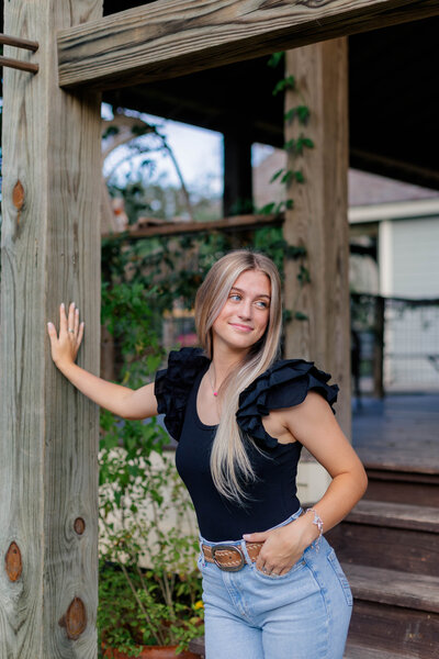 A blonde highschool senior with a black top and jeans on looks off into the distance while leaning against a wooden post in Alvin Texas