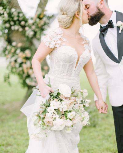A fine art image of a bride and groom kissing. The bride is wearing a gorgeous lace gown with a white and pink bouquet. The groom has on a white and black tuxedo