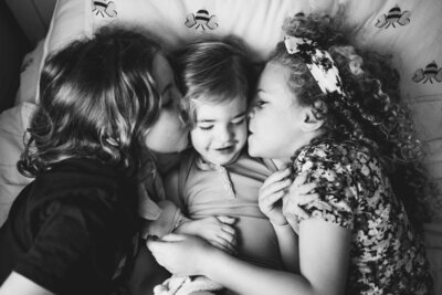 Black & white portrait of toddler boy is kissed by his two big sisters.