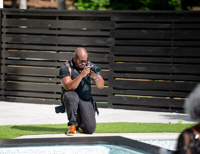 Professional wedding photographer kneels while taking photos outdoors with camera gear and equipment in natural setting.
