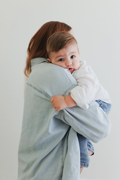 baby boy hugging his mama wearing jeans and white shirt while looking at camera