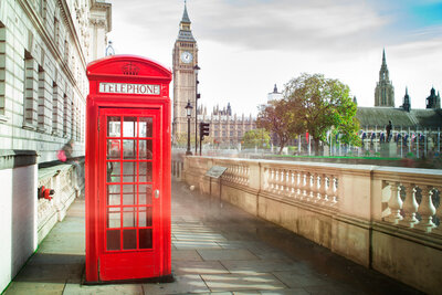 A classic red telephone box is positioned on a sidewalk in London. Big Ben and the Houses of Parliament are visible in the background under a partly cloudy sky. The scene combines iconic British architecture with a timeless symbol.