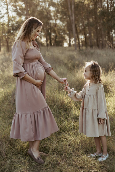 Pregnant mother in a pink dress cradling her bump and taking a flower from her six year old daughter in tan dress
