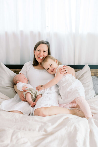A  new mom sits on her bed and snuggles her children while laughing at the camera of Northern Virginia Photographer