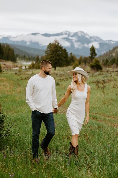 Couple walks together holding hands in a field with mountains in the background.