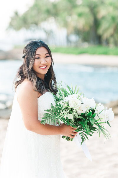 a bride standing on a beach behind the four seasons holding a bouquet with white florals and greenery