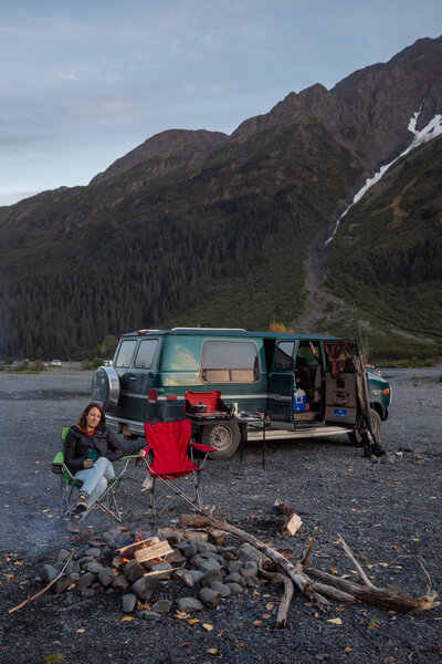 A green van is parked with it's door open as a woman sits in front of the van next to a campfire in Alaska.
