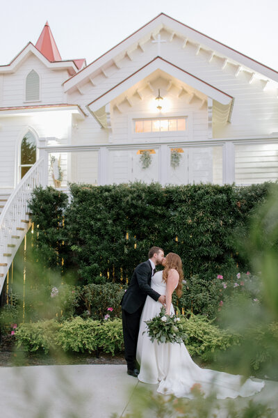 Chris in a black tux dips Ashlyn in her ivory bridal gown with cathedral train in front of the Tybee Island Chapel, a beautiful coastal savannah wedding venue. There are steps on their left leading up to the chapel and they are standing in front of green ivy and landscaping covering the lattice below.