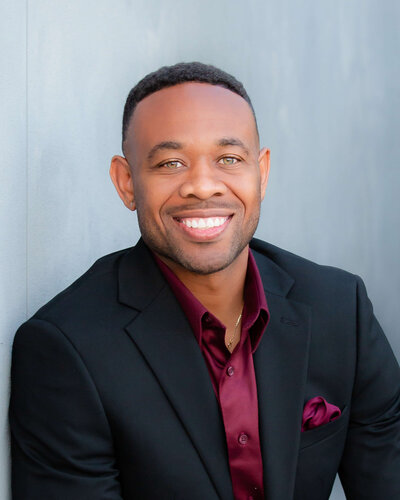 Professional headshot of a smiling man wearing a black suit with a burgundy dress shirt and pocket square, posed against a light gray background.
