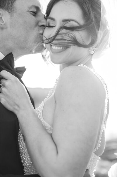 A smiling bride getting kisses under a veil underneath the sunlight by her husband by JoLynn Photography, a destination wedding photographer