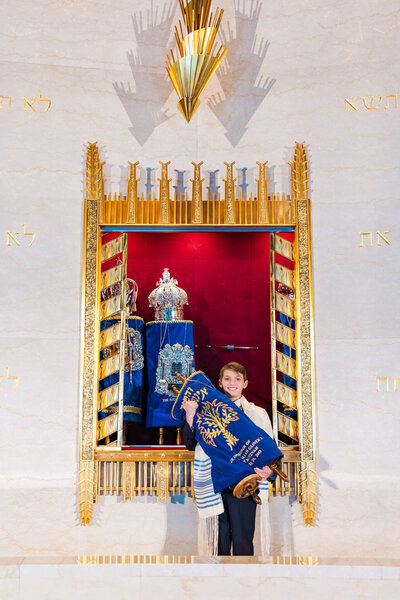A small teen boy holds a blue dressed torah in a black suit and tallit in a temple