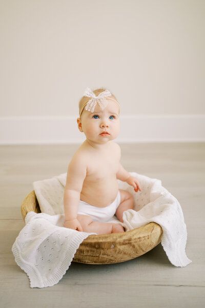Baby girl in white bow and white diaper cover sits in wooden bowl with white blanket during photography session in Raleigh NC