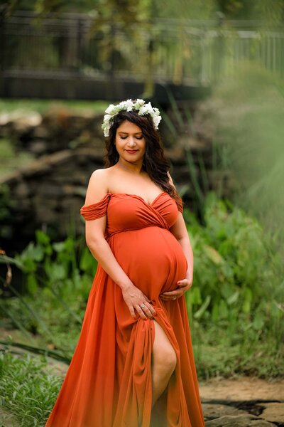 expectant charlotte mother in rust colored dress wearing a floral crown in a garden setting 