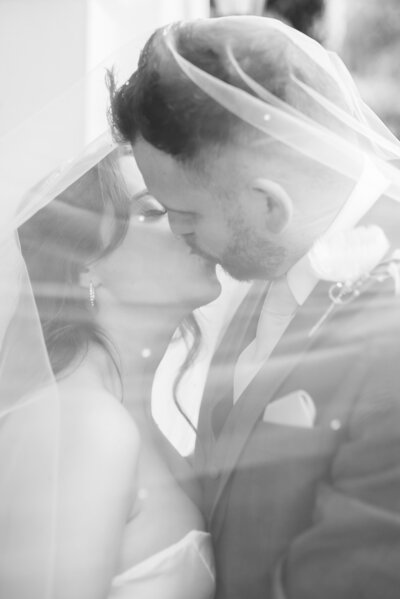 Black and white image of a bride and groom under the brides veil kissing.