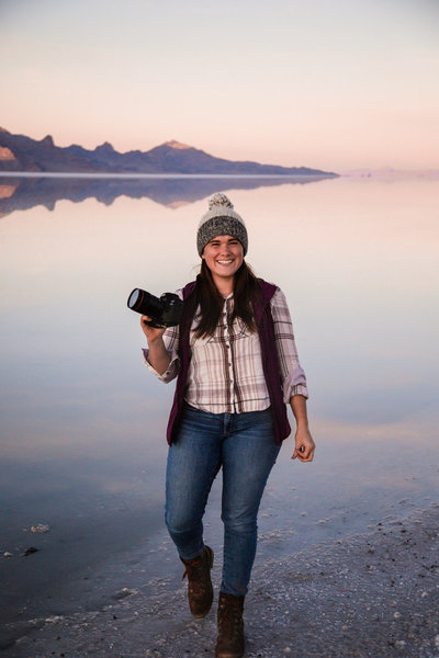 Photographer wearing a pink plaid shirt and jeans stands next to the Utah Salt Flats. The flats are covered with a thin layer of water making the surface reflect the sunset and mountain range in the distance.