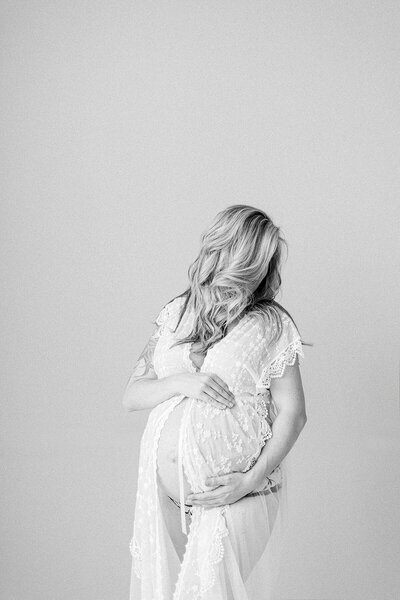 A black and white maternity portrait of a pregnant mom delicately holding her belly while looking over her shoulder in front of a window in a Dallas photography studio.