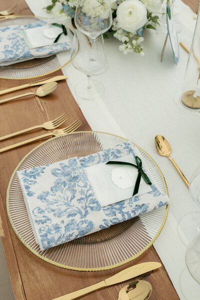 Elegant wedding table place setting featuring a watercolor menu wrapped in a soft fabric napkin, resting on a gold-rimmed glass charger. The setting is complemented by gleaming gold tableware and luxurious blue and white floral arrangements, adding a refined touch to the sophisticated decor.