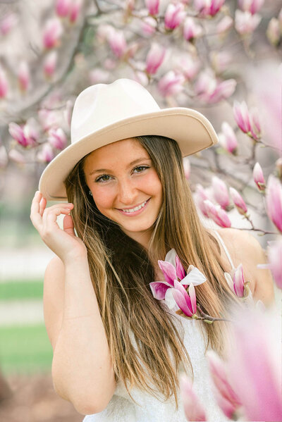 senior portrait of a blonde girl wearing a white hat standing next to pink magnolia flowers