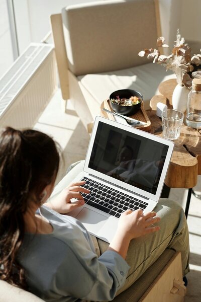 A woman sitting in a sunlit room, working on a laptop. She is wearing a light blue blouse and olive green pants, with her hair tied back. A small wooden side table beside her holds a glass of water, a bowl of salad, a white vase with dried flowers, and a water bottle. The space has a cozy, minimalist aesthetic with a neutral color palette.